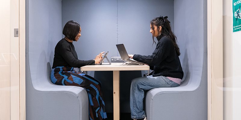Two students sat at table facing each other in library study pod